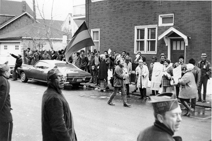 Black and white photograph of DRUM workers on strike. A man waves a mid the crowd along the street waves a flag.